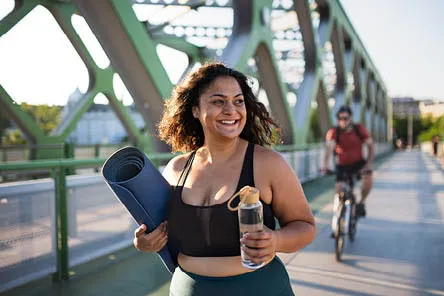 A woman walking over a bridge wearing gym clothing and carrying a yoga mat and water bottle