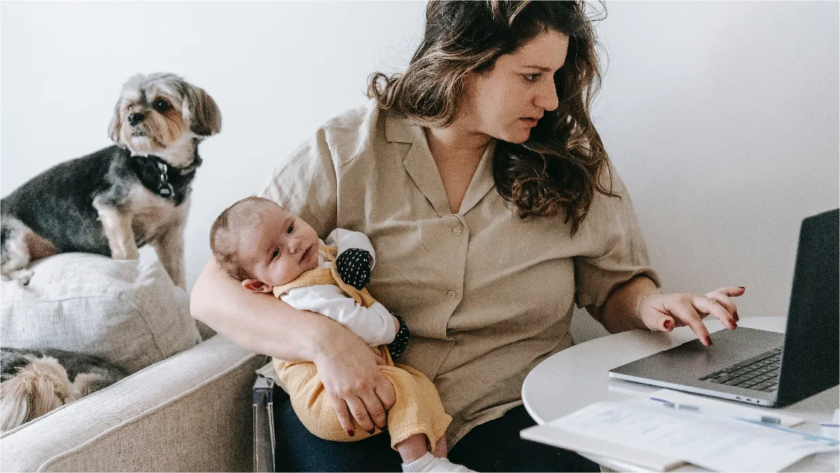 A new mother holds her baby in one arm as she works on her laptop with the other. Her two dogs watch her from the couch.